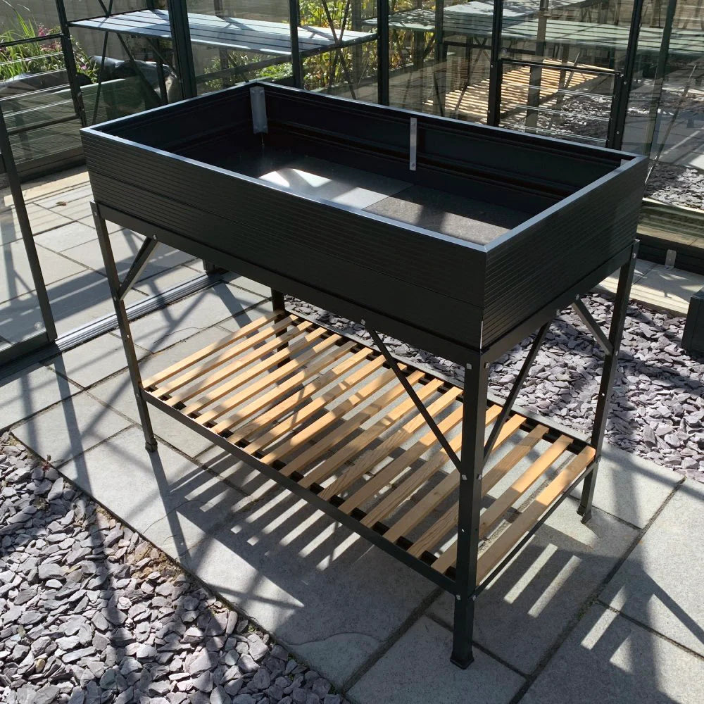 A black raised planter box stands on metal legs, with a wooden slatted shelf below. It is located inside a Rhino Greenhouse with gray slate flooring and glass-paneled walls.