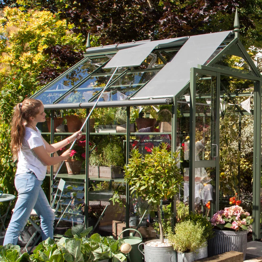Rhino Greenhouse with a glass structure is being adjusted by a person using a pole. It is surrounded by lush garden plants including flowers and vegetables, creating a vibrant outdoor setting.