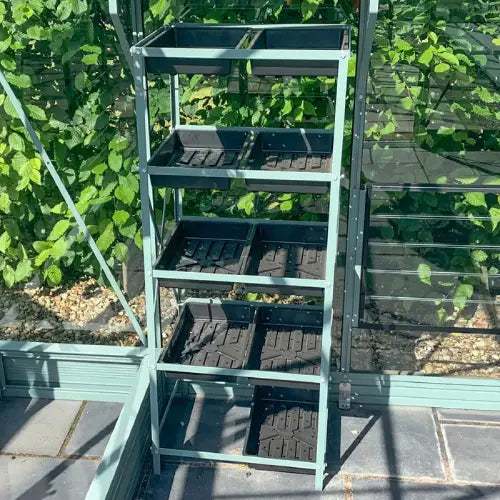 A green metal shelving unit holds several black planting trays, standing inside a Rhino Greenhouse. Sunlight filters in, and lush green plants grow in the background.