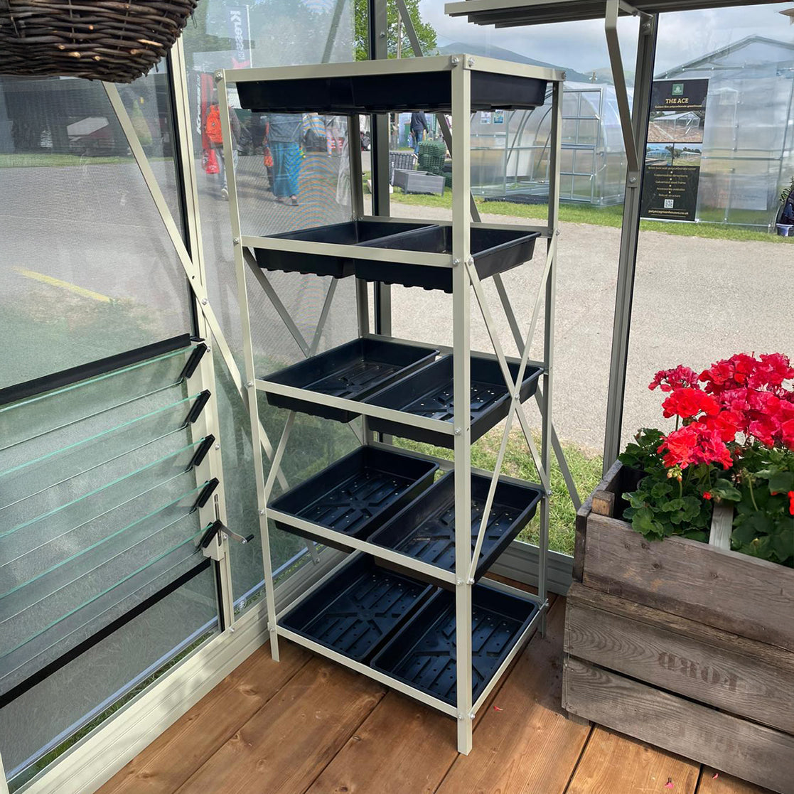 A metal shelving unit with black trays stands in a Rhino Greenhouse on a wooden floor. Nearby, there's a planter with red flowers, and outside, blurred figures and more Rhino Greenhouses are visible.