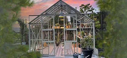 A glass Rhino Greenhouse filled with plants and gardening tools stands in a garden. The sky is vibrant with a sunset in the background, surrounded by greenery.