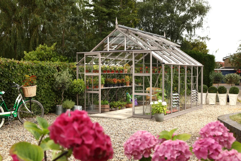 A glass Rhino Greenhouse stands in a gravel garden, containing vibrant potted plants. Nearby, a bicycle with a basket leans against a hedge, surrounded by lush greenery and pink flowers.
