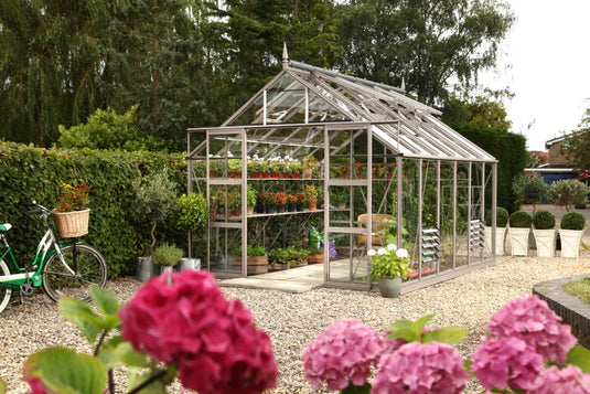 A glass Rhino Greenhouse stands in a gravel garden, containing vibrant potted plants. Nearby, a bicycle with a basket leans against a hedge, surrounded by lush greenery and pink flowers.