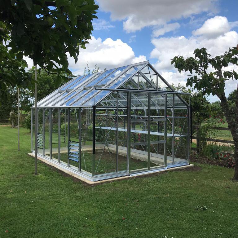 A glass Rhino Greenhouse with a peaked roof stands on lush grass. It's surrounded by trees and garden paths, with a partly cloudy sky above.