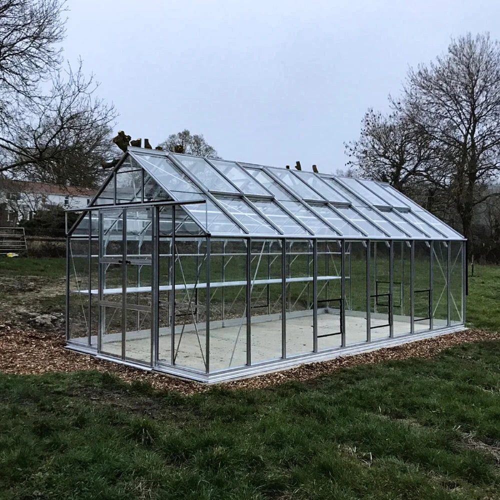 A glass Rhino Greenhouse with a metal frame stands on a grassy field, surrounded by trees, under an overcast sky.