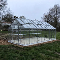 A glass Rhino Greenhouse with a metal frame stands on a grassy field, surrounded by trees, under an overcast sky.
