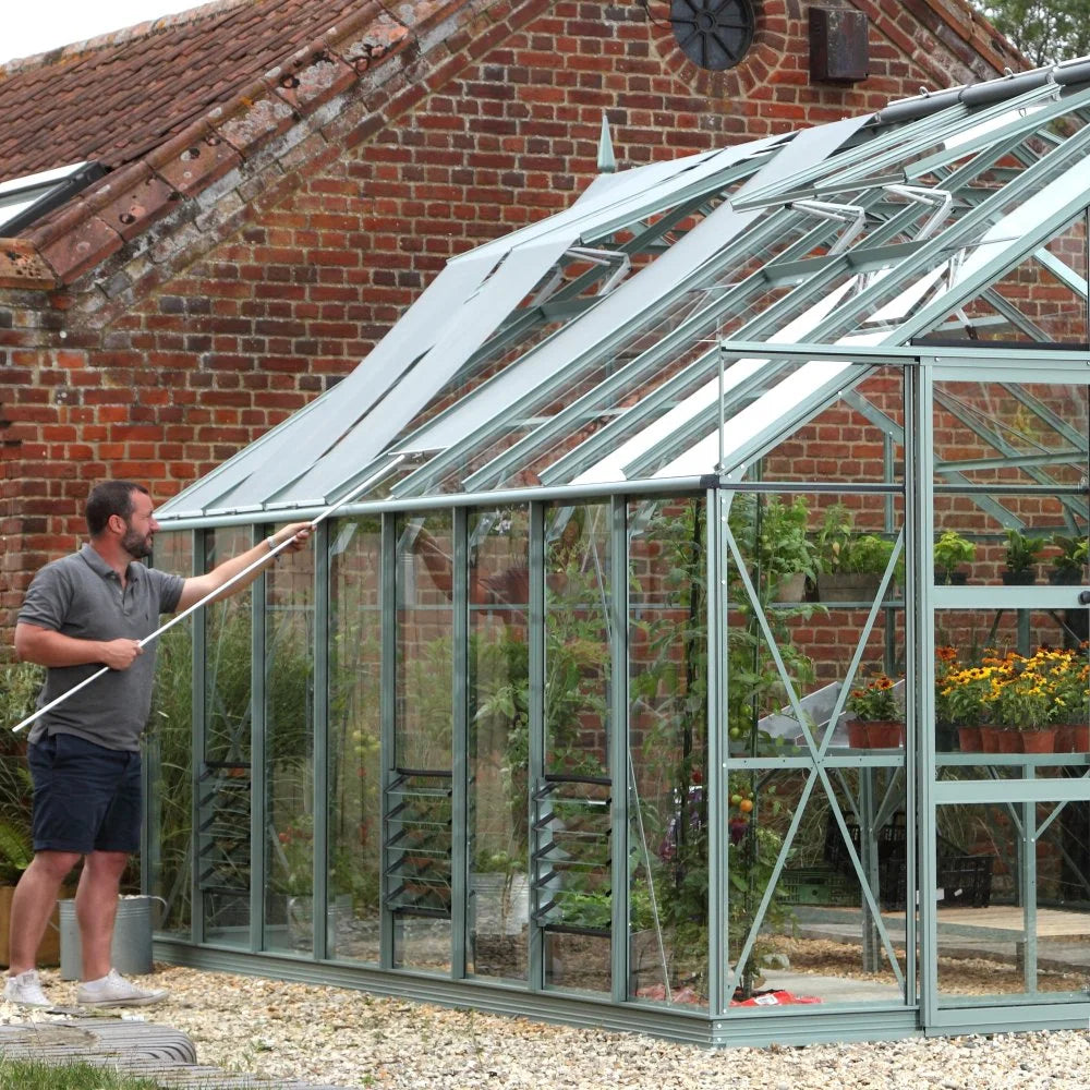 A man cleans the glass panels of a large, modern Rhino Greenhouse with a tool, situated beside a rustic brick building, surrounded by potted plants and gravel.