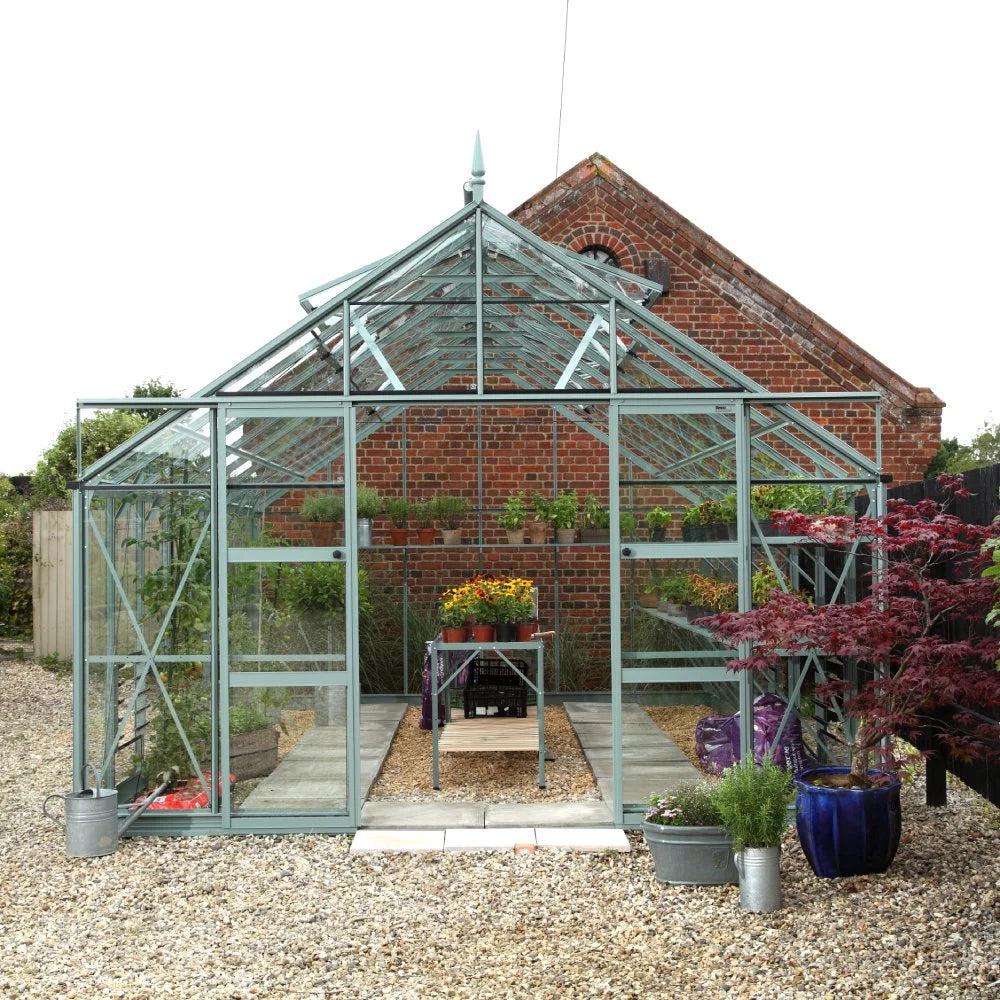 A glass Rhino Greenhouse showcases potted plants and flowers inside, surrounded by a gravel path. It is positioned in front of a brick building, with additional pots on either side.