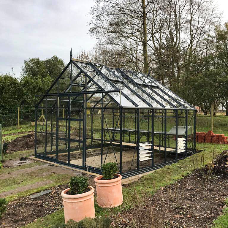 A glass-paneled Rhino Greenhouse stands in a garden, with potted plants nearby and surrounded by trees and a lawn, suggesting a tranquil outdoor setting.