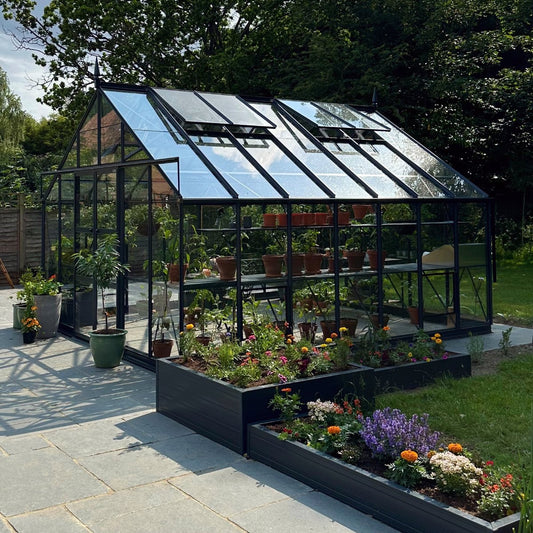 A glass Rhino Greenhouse shelters potted plants, reflecting sunlight, surrounded by blooming flowers in raised beds, with a wooden fence and green trees in the background.