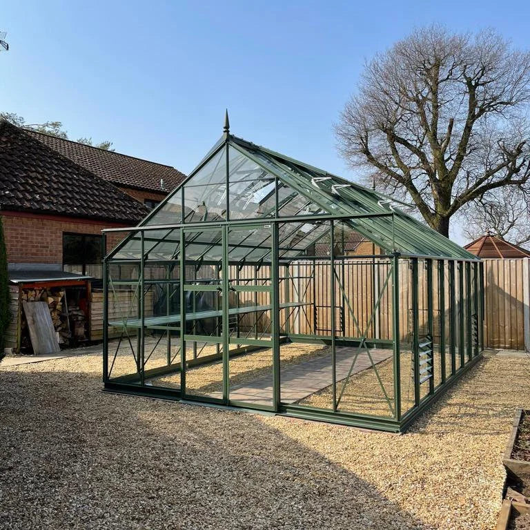A large, green-framed Rhino Greenhouse stands on a gravel area, adjacent to a brick building and wooden fence, with bare trees visible under a clear blue sky.