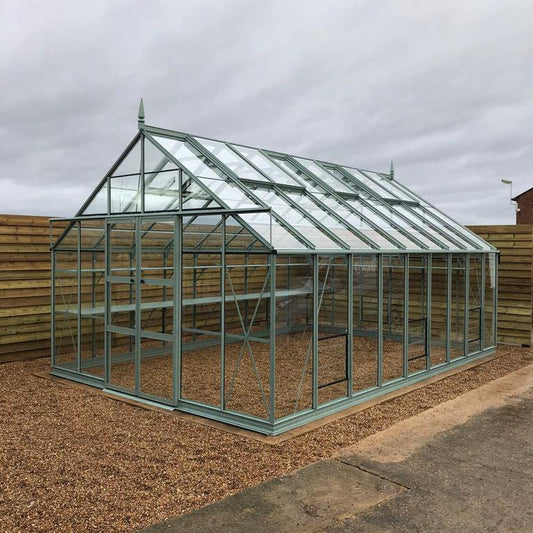 A glass and metal Rhino Greenhouse stands on a gravel surface, surrounded by wooden fencing, under an overcast sky.