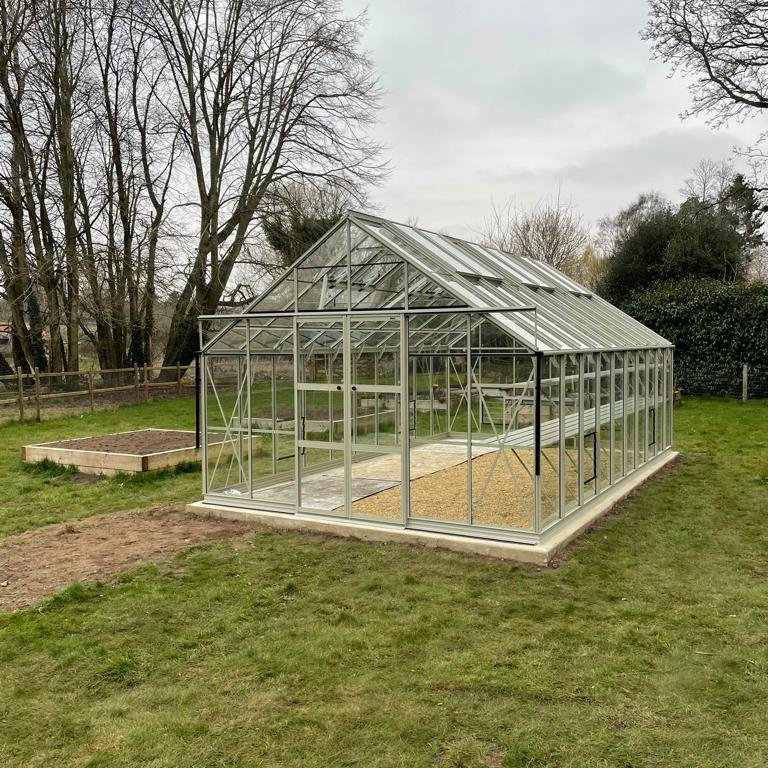 A large glass Rhino Greenhouse stands in a grassy garden, with metal framing and a gravel pathway inside. Surrounding the Rhino Greenhouse are trees and wooden planters in the background.