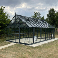 Glass Rhino Greenhouse with a pointed roof stands on a concrete slab, surrounded by grassy field and trees under a cloudy sky.
