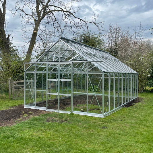 A Rhino Greenhouse stands in a grassy garden, surrounded by trees. It has a metal frame and glass panels, allowing natural light to enter, suitable for growing plants and vegetables.