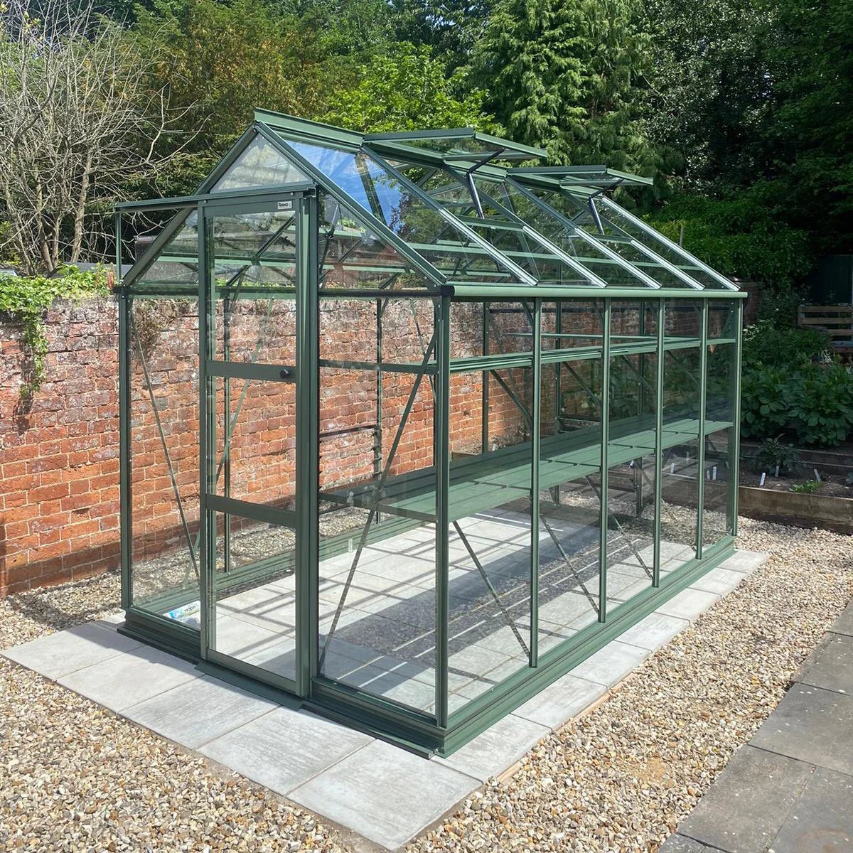 A green-framed glass Rhino Greenhouse stands on a paved area, surrounded by gravel, in a garden with brick walls and lush greenery in the background.