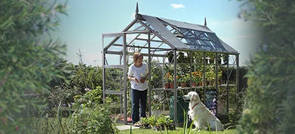 A Rhino Greenhouse stands in a garden, with plants inside. A person tends to the plants, while a dog sits on the grass nearby. Lush foliage surrounds the scene.