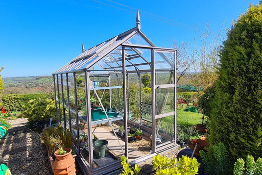 A glass Rhino Greenhouse stands amid a lush garden landscape. It features triangular roof panels, open door, and potted plants inside and around, surrounded by trees and expansive countryside views.