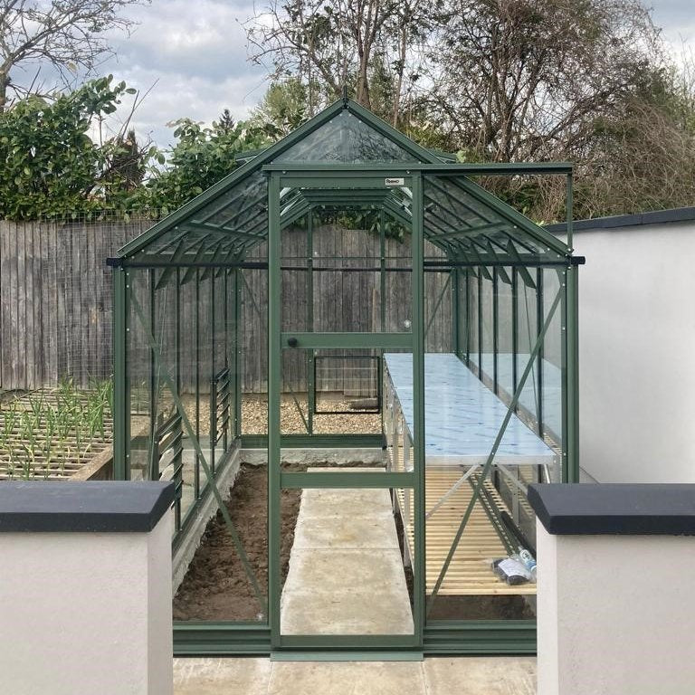 A green metal-frame Rhino Greenhouse stands on a paved path, with shelving inside. It is surrounded by a wooden fence and garden with trees. The text reads: Rhino.
