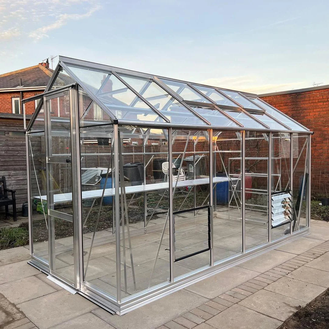 A glass and aluminum-framed Rhino Greenhouse stands on a paved patio, surrounded by garden structures and a red brick wall in a residential backyard setting.
