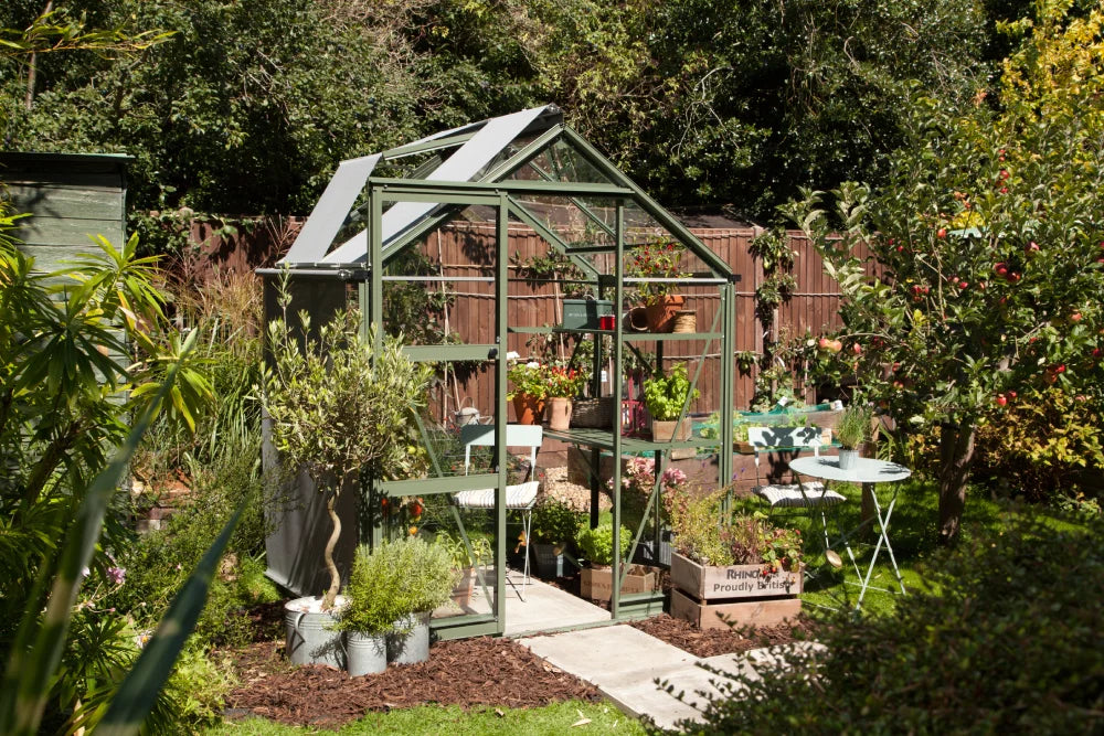 A green Rhino Greenhouse stands in a lush garden, filled with potted plants. Nearby, a small table and chair set complements the greenery. A wooden crate reads, Rhino, Proudly British.