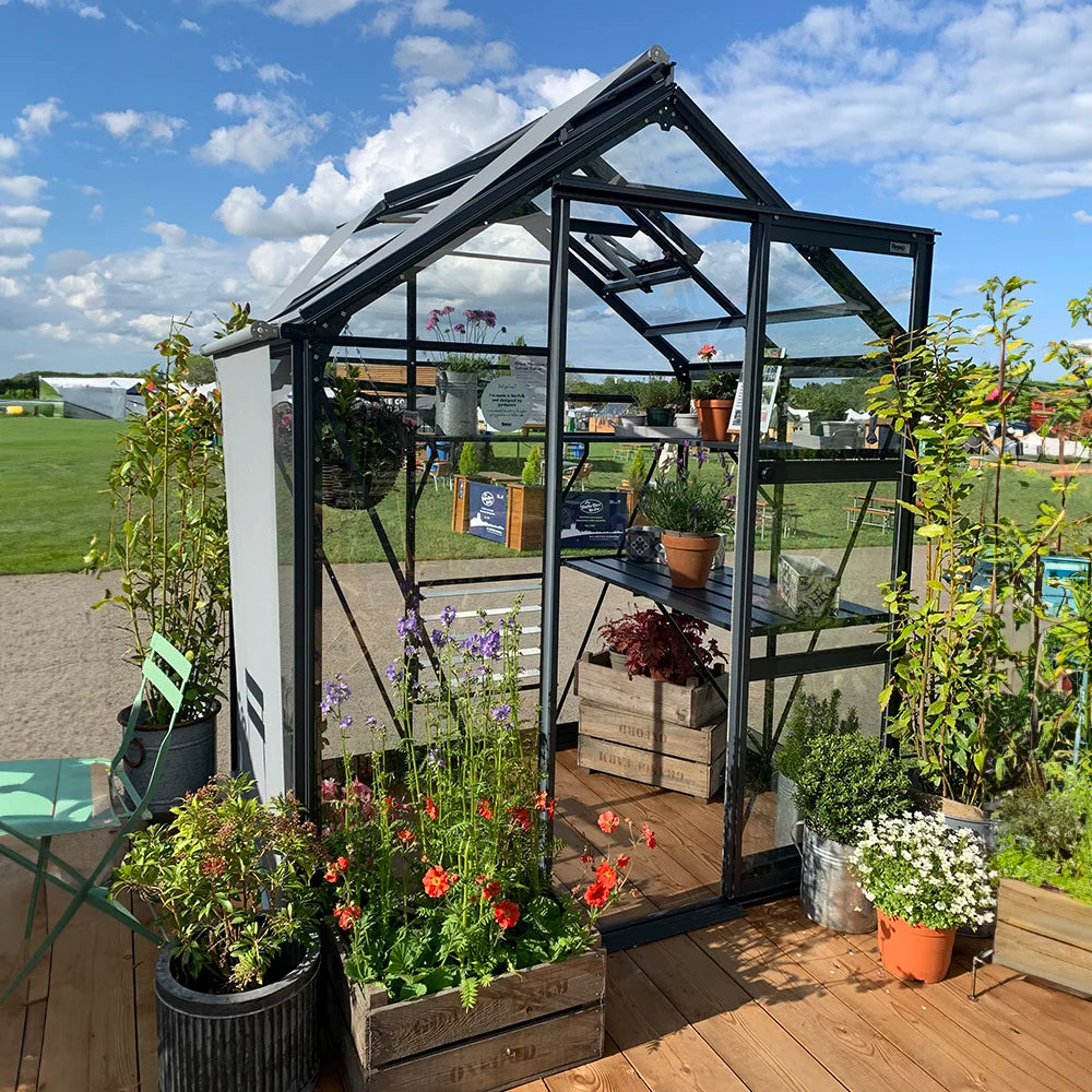 A glass Rhino Greenhouse stands with various plants inside and outside in pots and crates. It is set on a wooden deck with a grassy field and clear blue sky in the background.