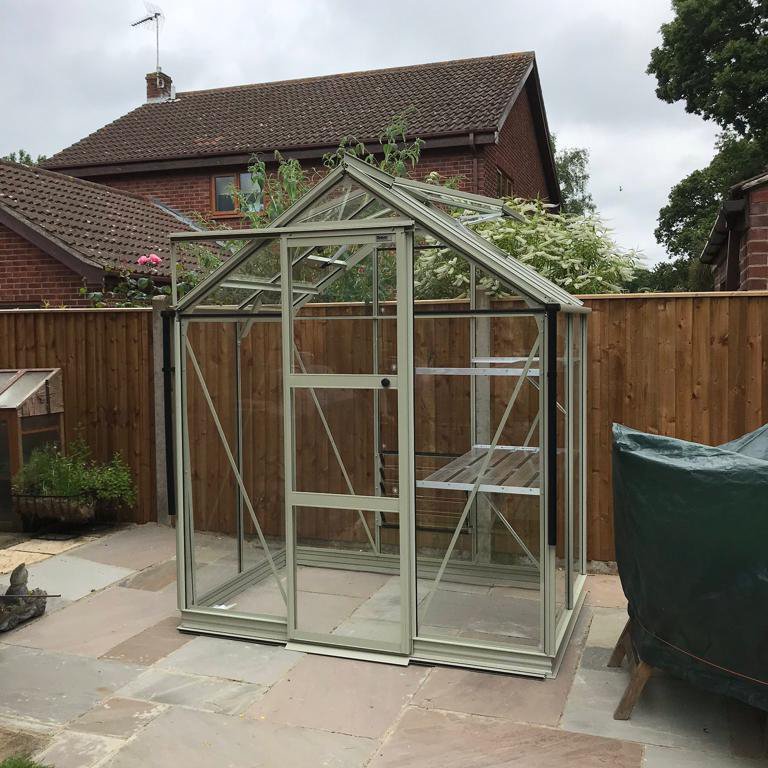 A small, glass-panel Rhino Greenhouse stands on a paved patio, surrounded by wooden fencing and adjacent to a residential brick house.