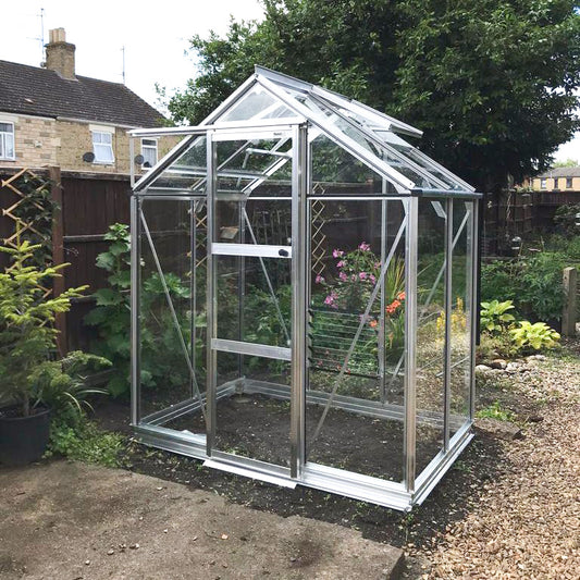 A glass and metal Rhino Greenhouse stands in a garden, surrounded by green foliage and adjacent to a house. Inside, flowers and plants grow, receiving ample sunlight through transparent panels.
