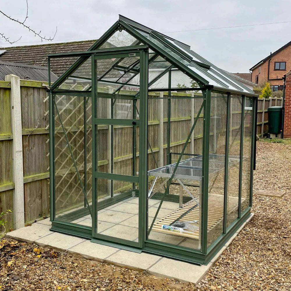 Rhino Greenhouse with a metal frame and glass panels stands on paving stones in a gravel backyard. A bench and small gardening tools are inside. Nearby fences and houses provide context.