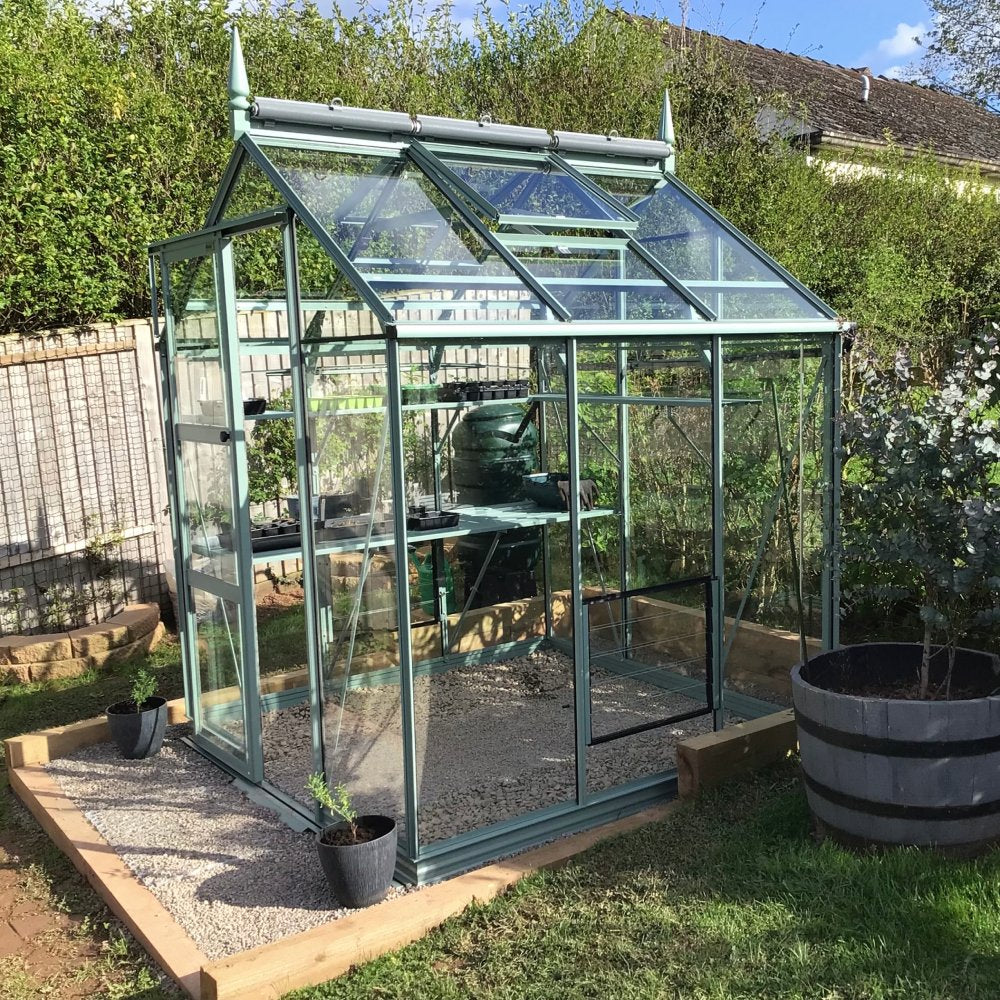 A glass Rhino Greenhouse stands on a gravel foundation, containing various gardening tools and plants. It is surrounded by grass, hedges, and a wooden fence, under a clear blue sky.