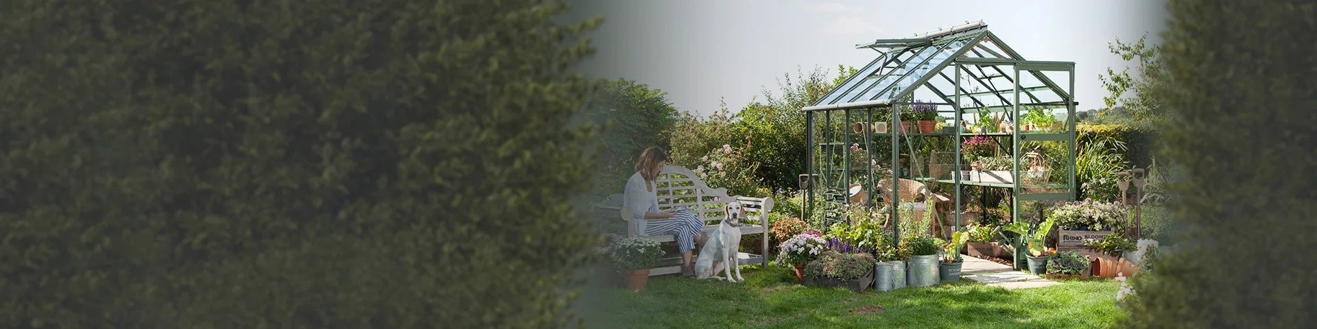 A glass Rhino Greenhouse filled with plants stands in a lush garden. Nearby, a person sits on a bench beside a dog, surrounded by potted flowers and gardening tools.