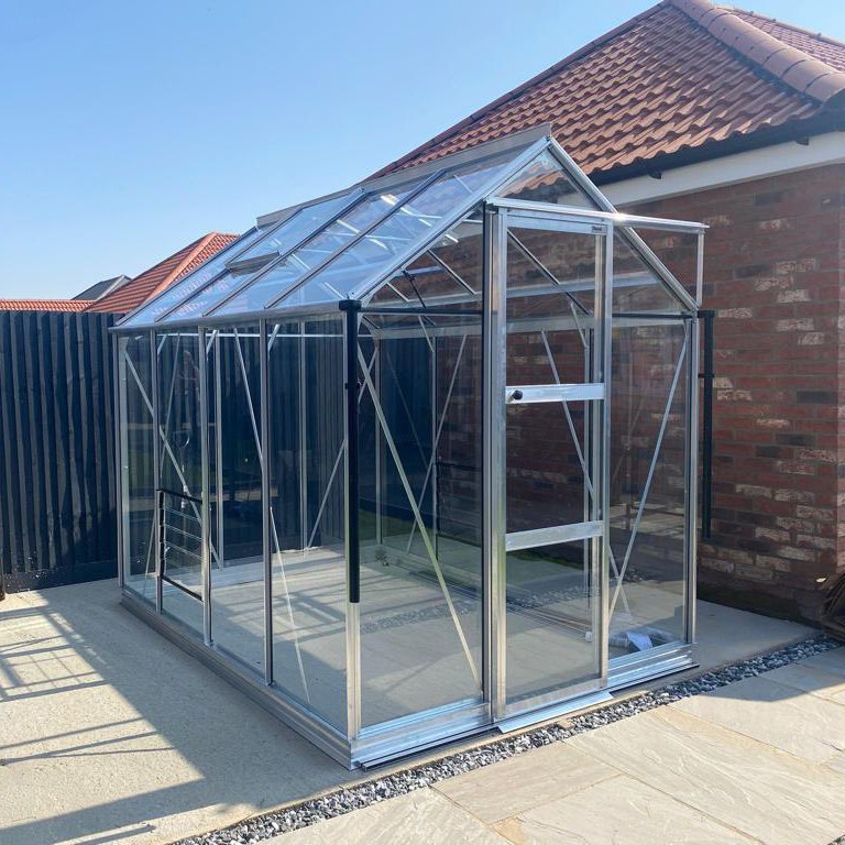 A glass Rhino Greenhouse with metal framing stands on a stone patio beside a brick wall and a wooden fence under a clear blue sky.