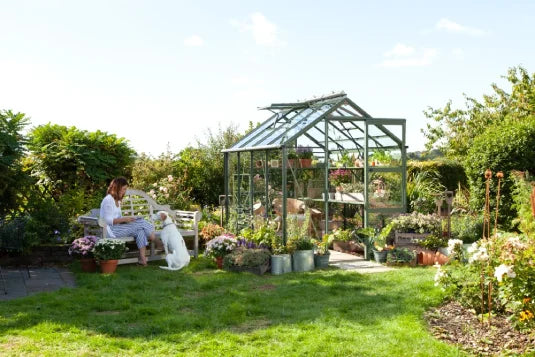 A glass Rhino Greenhouse stands in a garden, surrounded by potted plants and flowers. A person sits on a bench with a dog nearby, enjoying the sunny, lush environment.