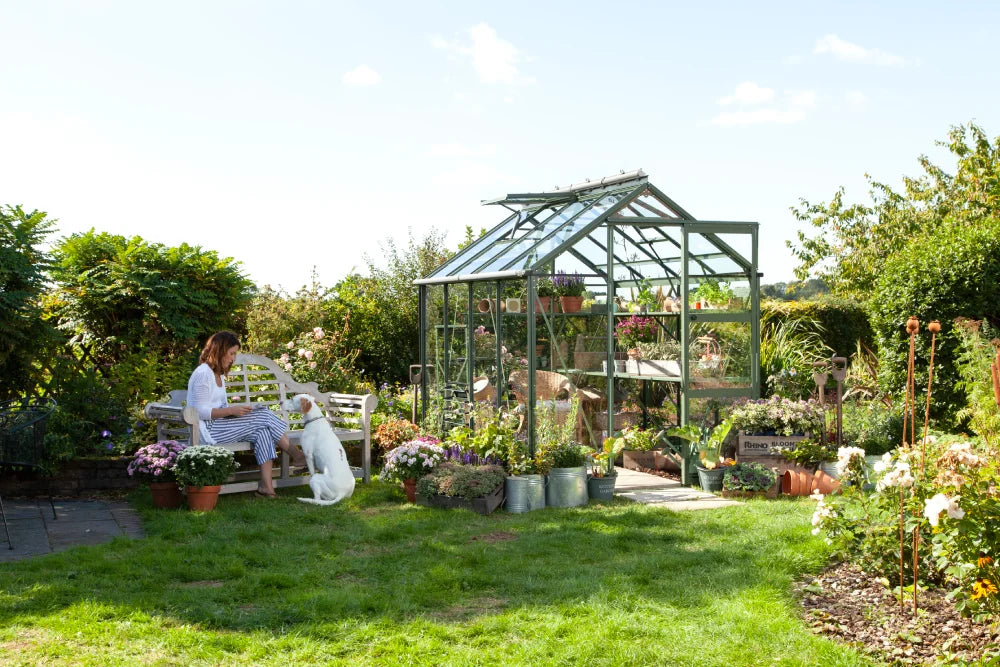Rhino Greenhouse with glass panels stands in lush garden, surrounded by potted flowers. Nearby, a woman in striped clothing sits on a bench, interacting with a white dog amidst vibrant greenery.