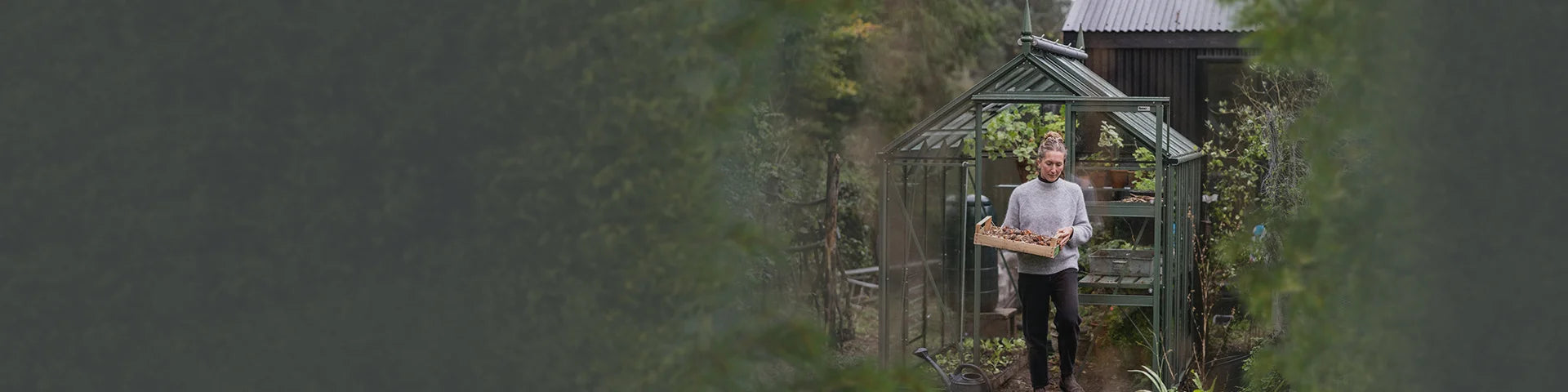 A person carries a crate of plants outside a green Rhino Greenhouse surrounded by lush garden foliage and rustic structures.