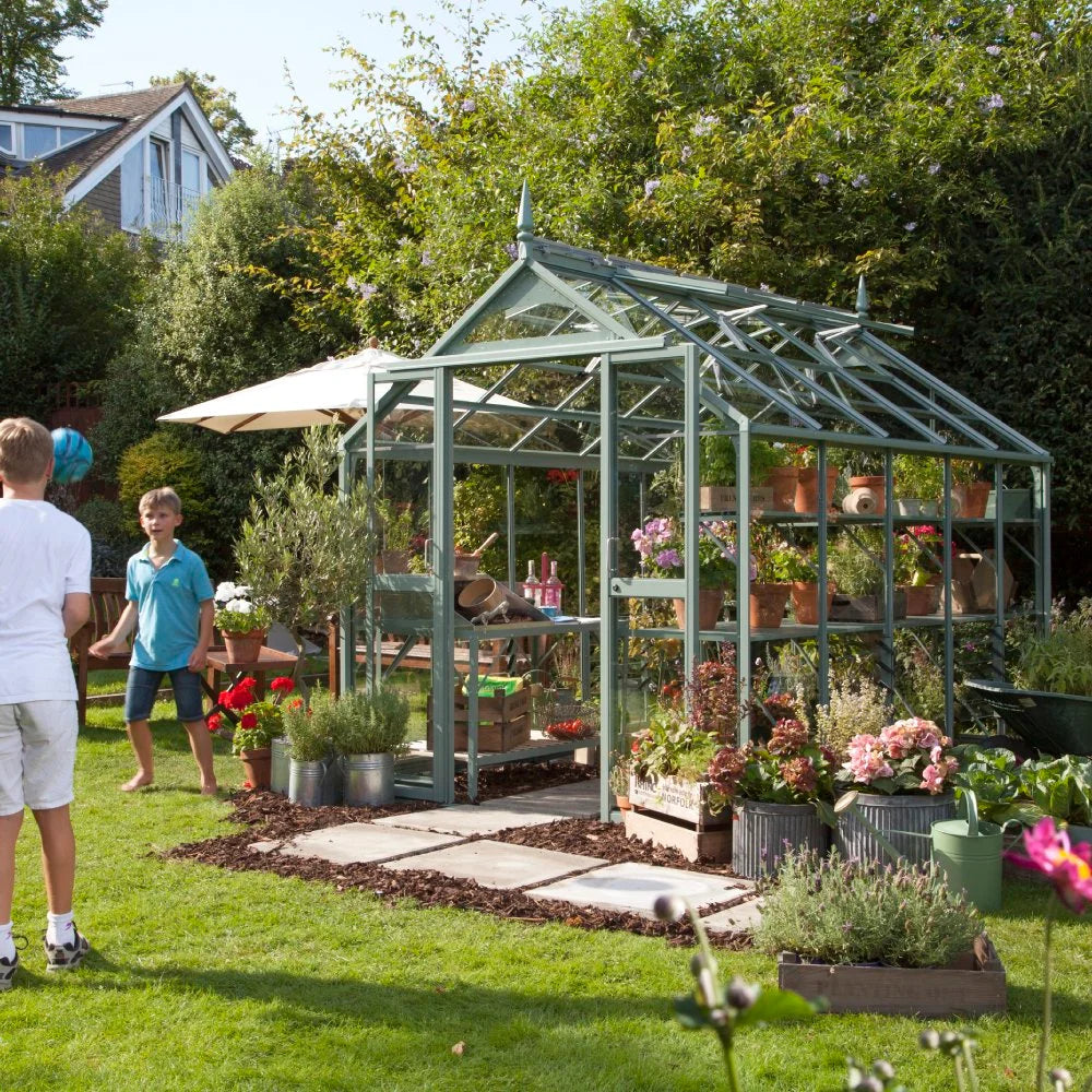 A glass Rhino Greenhouse filled with potted plants stands in a lush garden. Two children play with a ball nearby, and various gardening tools are scattered around.