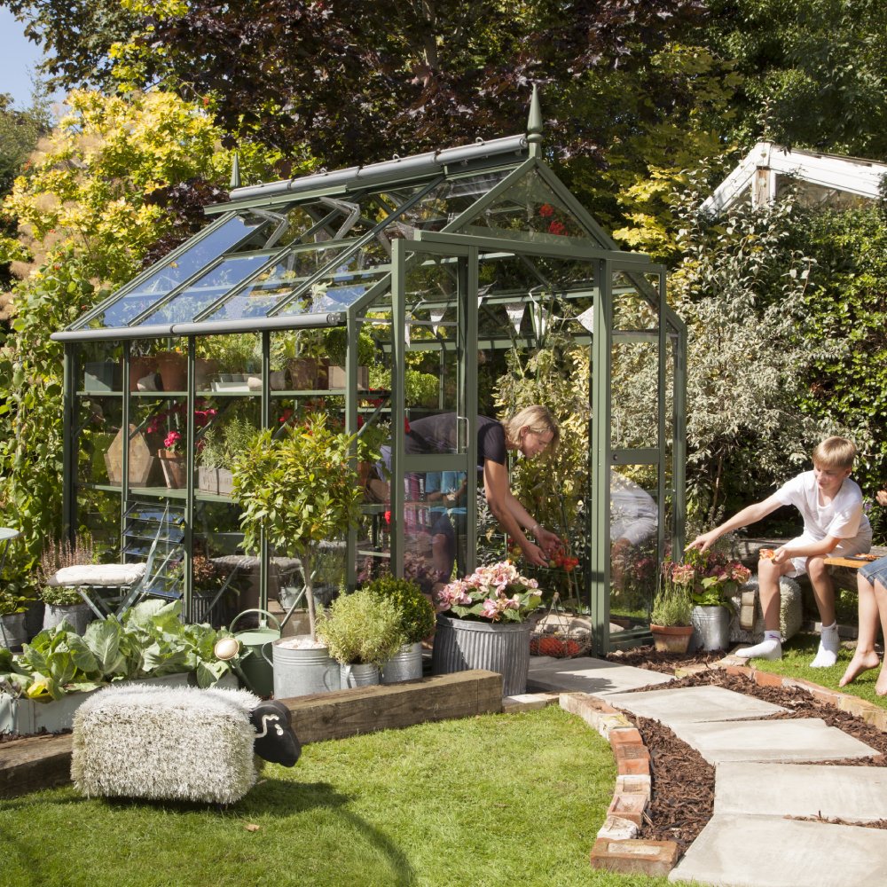 A green metal-framed Rhino Greenhouse shelters various plants. A person tends to flowers inside, while a nearby child points at other plants. It's surrounded by a lush garden and paved walkway.