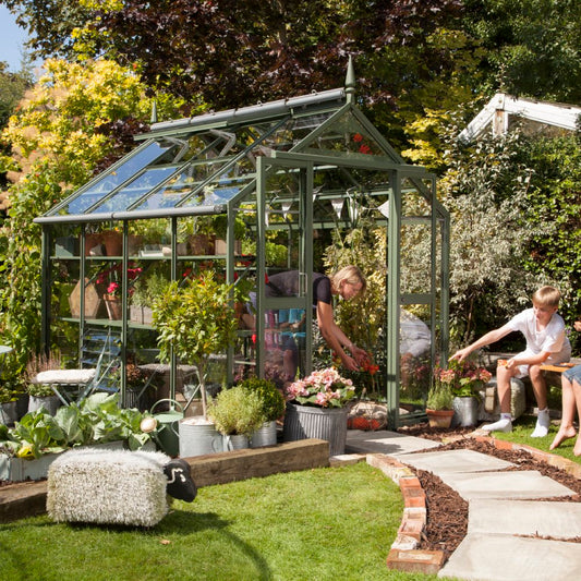 A green metal-framed Rhino Greenhouse shelters various plants. A person tends to flowers inside, while a nearby child points at other plants. It's surrounded by a lush garden and paved walkway.