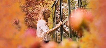 A person is interacting with a glass-paneled structure, possibly a Rhino Greenhouse. They are surrounded by vibrant flowers in a garden setting, emphasizing a connection with nature.