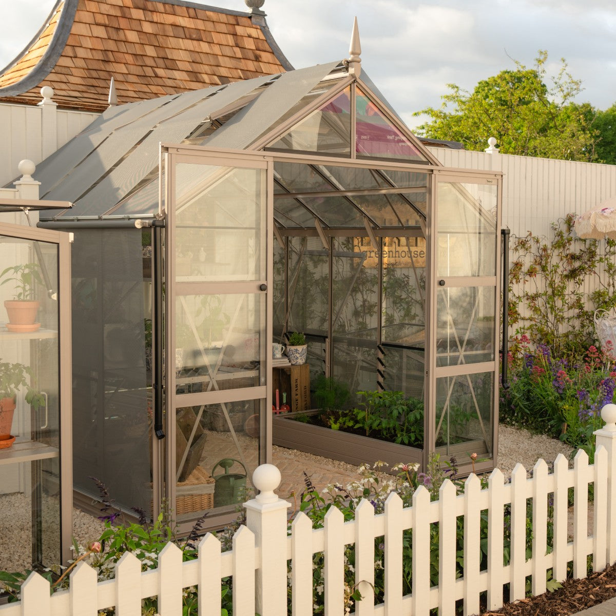 A glass Rhino Greenhouse with open doors displays potted plants inside. It's surrounded by a white picket fence, flowering plants, and a wooden shed in a garden setting.
