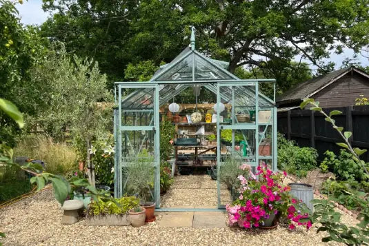 A glass Rhino Greenhouse stands in a garden, filled with potted plants and hanging lanterns, surrounded by gravel paths and blooming flowers, under a canopy of mature trees.