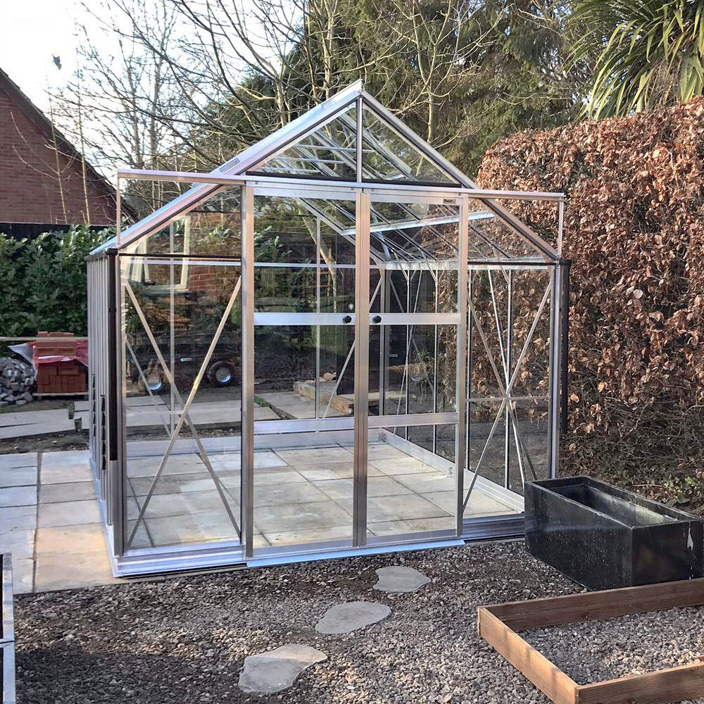 A glass-paneled Rhino Greenhouse stands on concrete slabs, surrounded by a garden with bare trees and hedges. Nearby, a rectangular black planter and small stepping stones are visible.