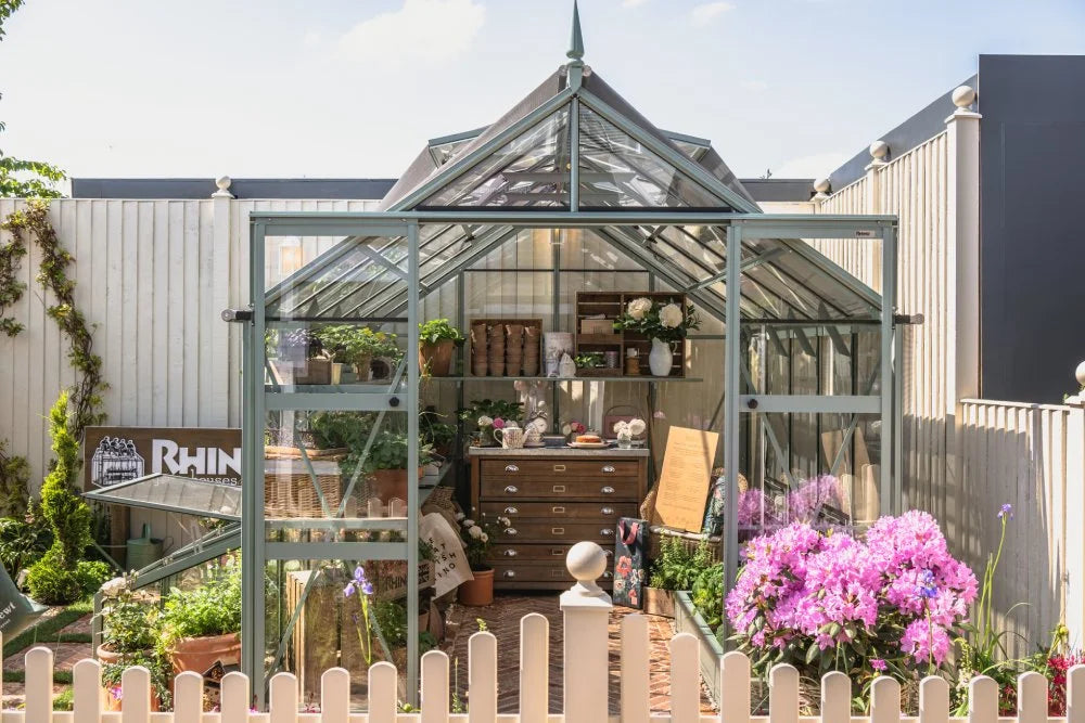A transparent Rhino Greenhouse displays various plants and gardening tools. It is surrounded by a picket fence and vibrant flowers. A sign reads Rhino Rhino Greenhouses against a wooden backdrop.
