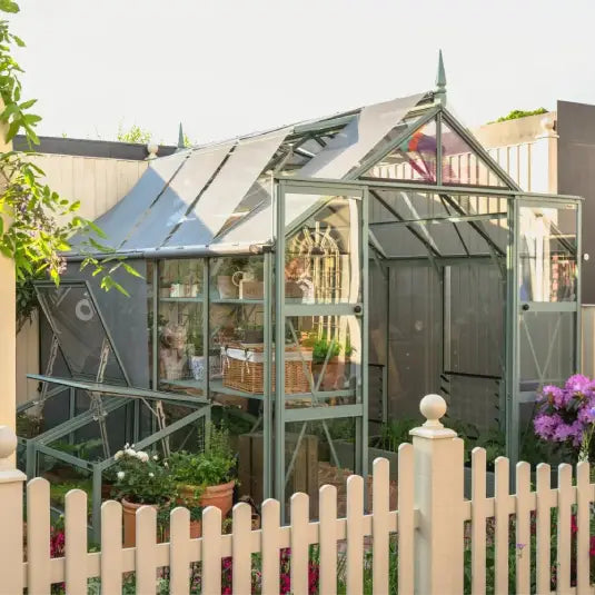 A glass Rhino Greenhouse with a pointed roof houses potted plants and gardening tools. It stands in a garden, enclosed by a white picket fence, with flowers and greenery surrounding it.