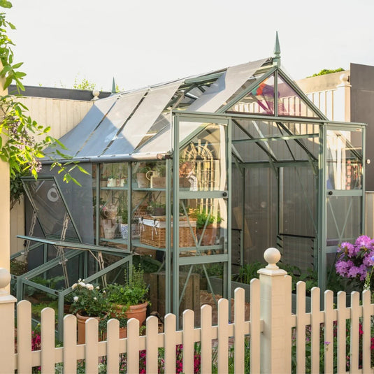 A medium-sized glass Rhino Greenhouse stands in a garden behind a white picket fence, surrounded by potted plants and flowers. It features a sloped roof and houses various gardening tools and plants.
