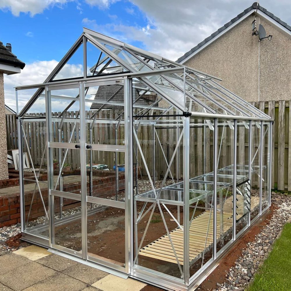 Glass Rhino Greenhouse with metal frame stands on a garden path, surrounded by a wooden fence, adjacent to a house. Clear skies overhead. No text visible.
