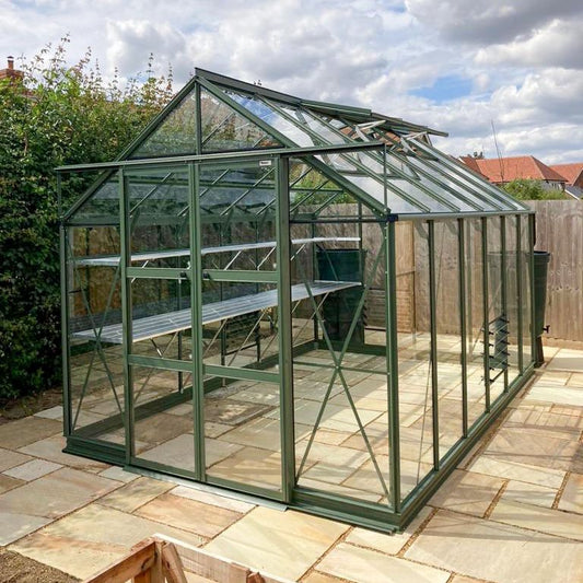 A green-framed glass Rhino Greenhouse, with open roof vents, stands on a stone patio in a garden surrounded by wooden fences and lush greenery under a cloudy sky.