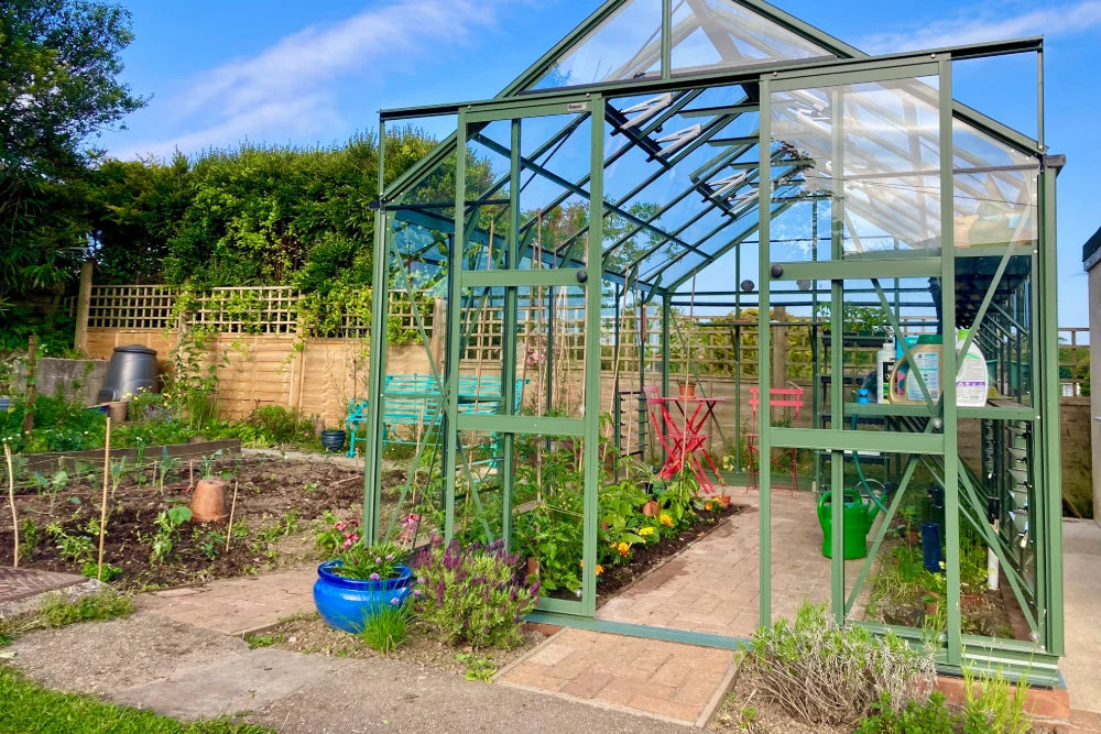 A green, glass-paneled Rhino Greenhouse stands in a garden. Inside, plants grow alongside gardening tools and accessories. Outside, a garden area with potted plants and fencing is visible.