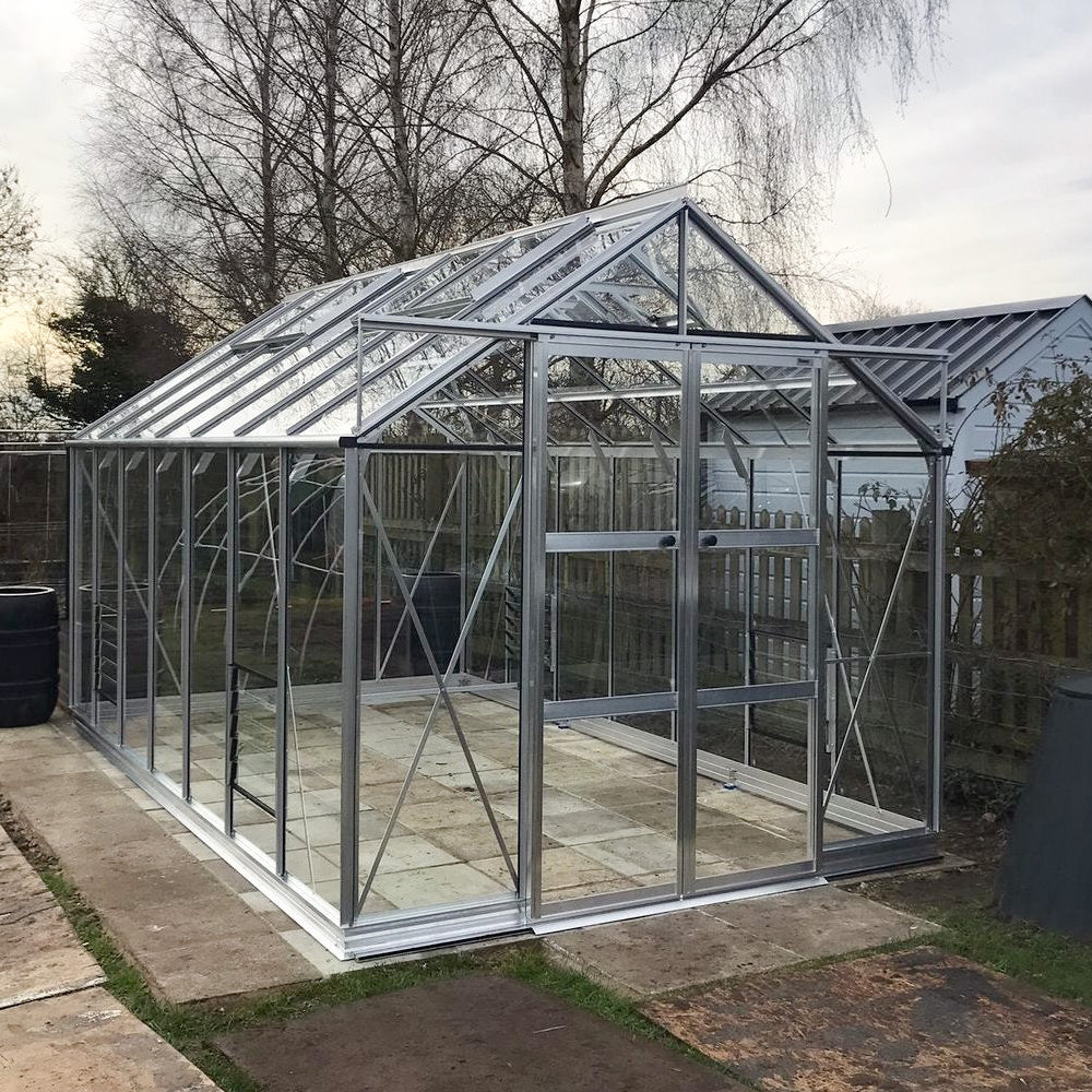A large, transparent Rhino Greenhouse made of glass and metal sits on a paved area. It has a pitched roof and is surrounded by bare trees and a wooden fence.