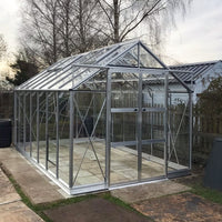 A large, transparent Rhino Greenhouse made of glass and metal sits on a paved area. It has a pitched roof and is surrounded by bare trees and a wooden fence.
